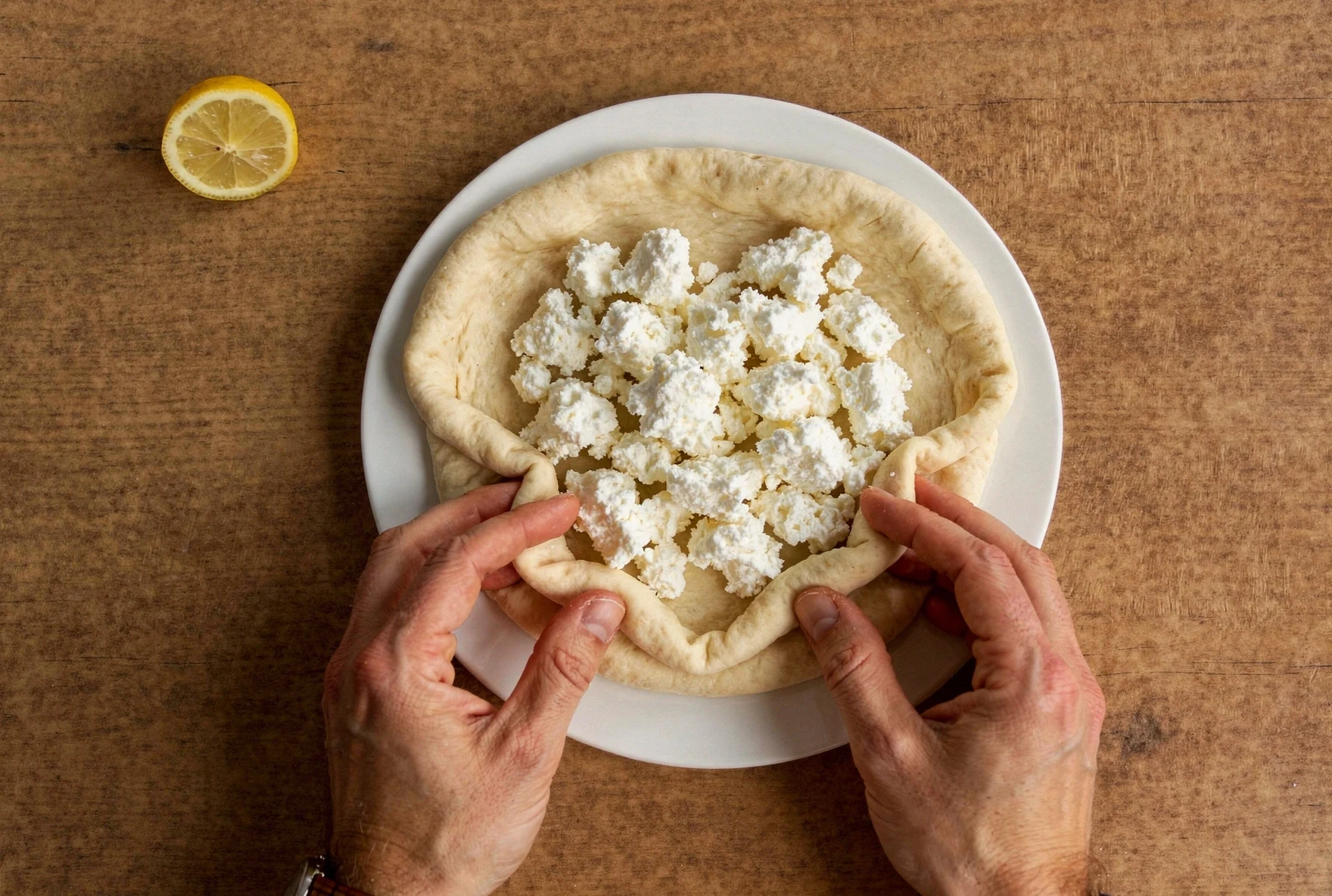 Hands folding khachapuri dough over cheese filling on a floured wooden surface